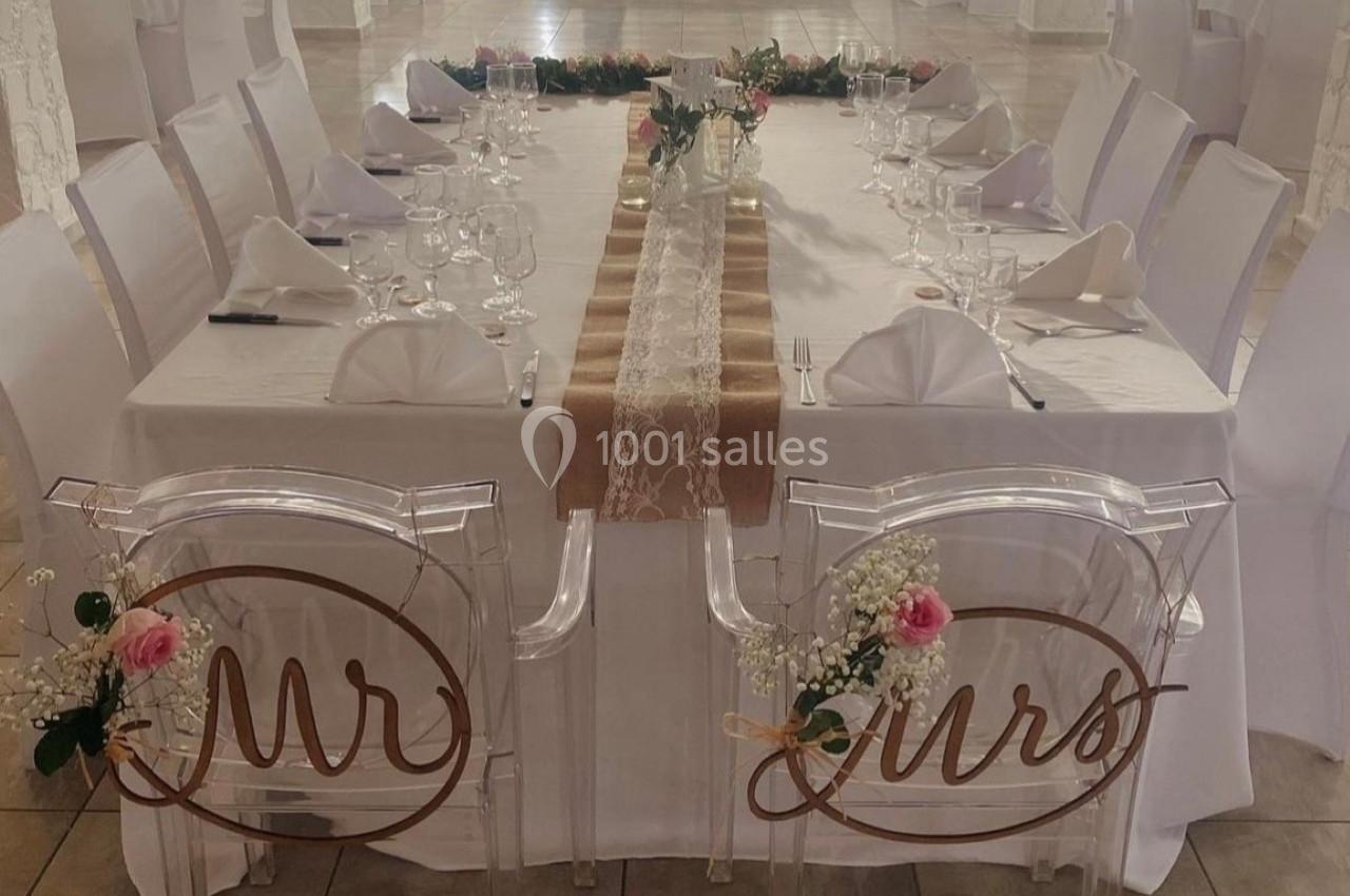 Table de mariage décorée avec nappes blanches, chemins de table en dentelle et chaises transparentes ornées des mots ’Mr’ et…