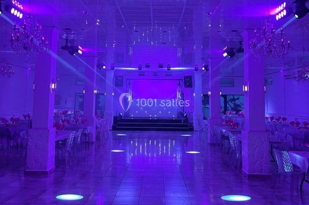Salle de réception éclairée en violet avec tables dressées, lustres suspendus et une scène au fond.