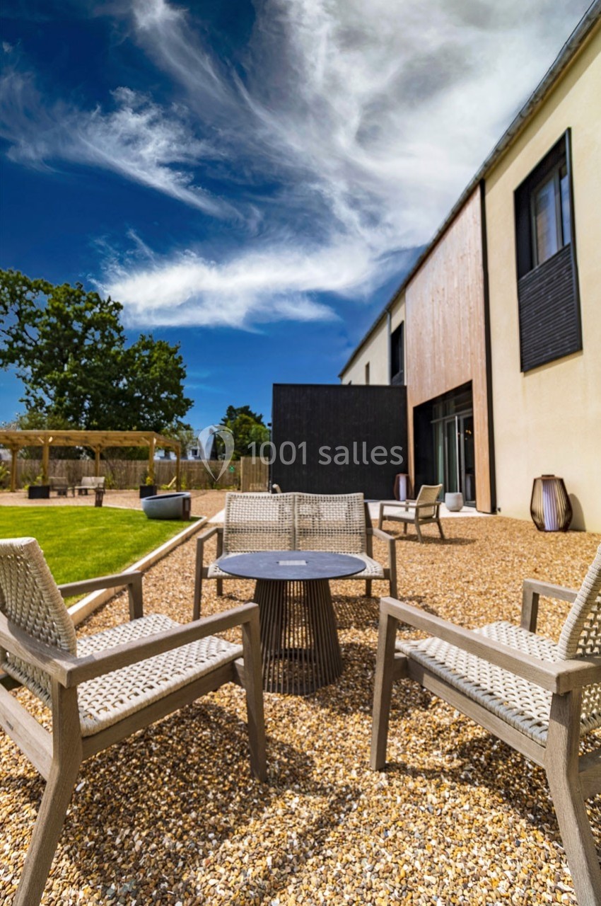 Terrasse avec mobilier de jardin en bois et métal sur gravier, entourée de pelouse et d'un bâtiment moderne.