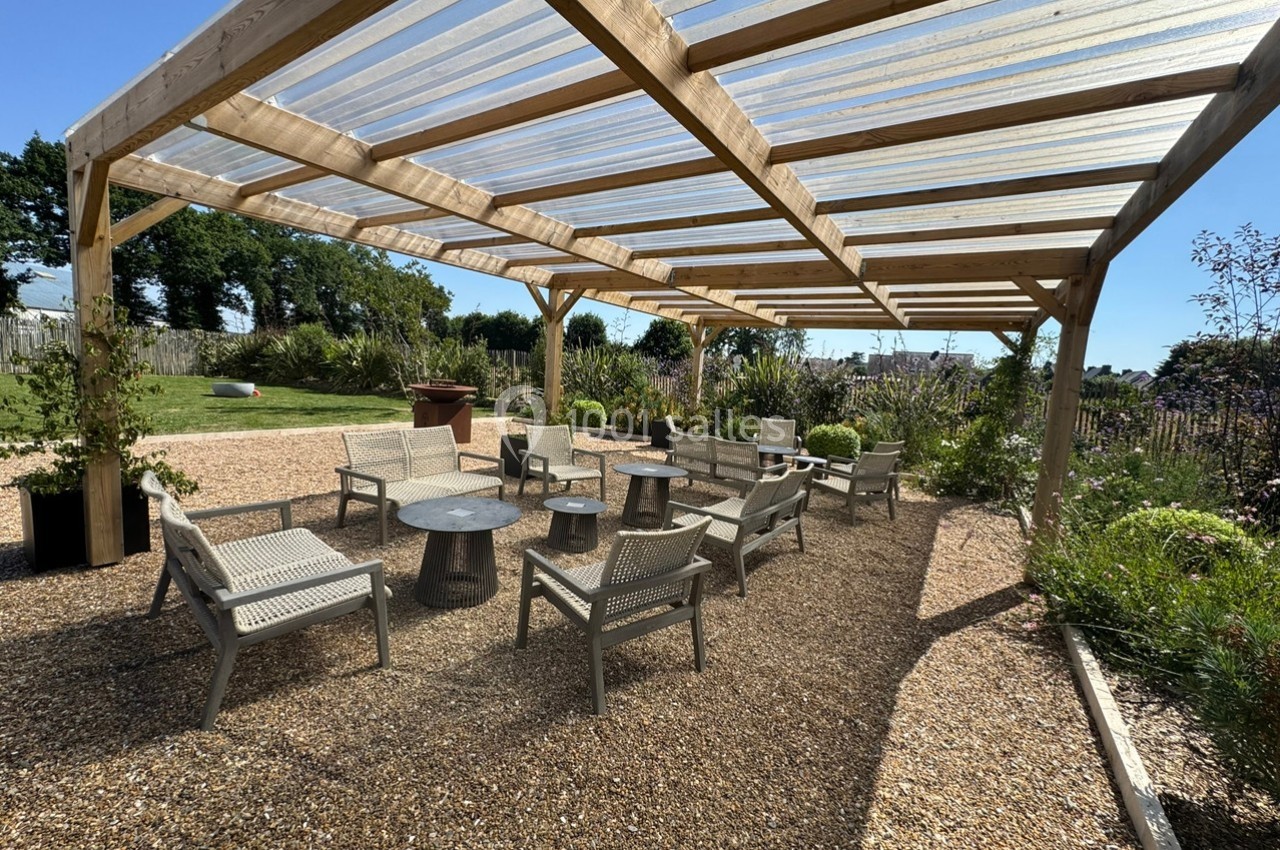 Pergola en bois avec toit transparent, abritant des tables et chaises sur un sol en gravier, entourée de végétation.