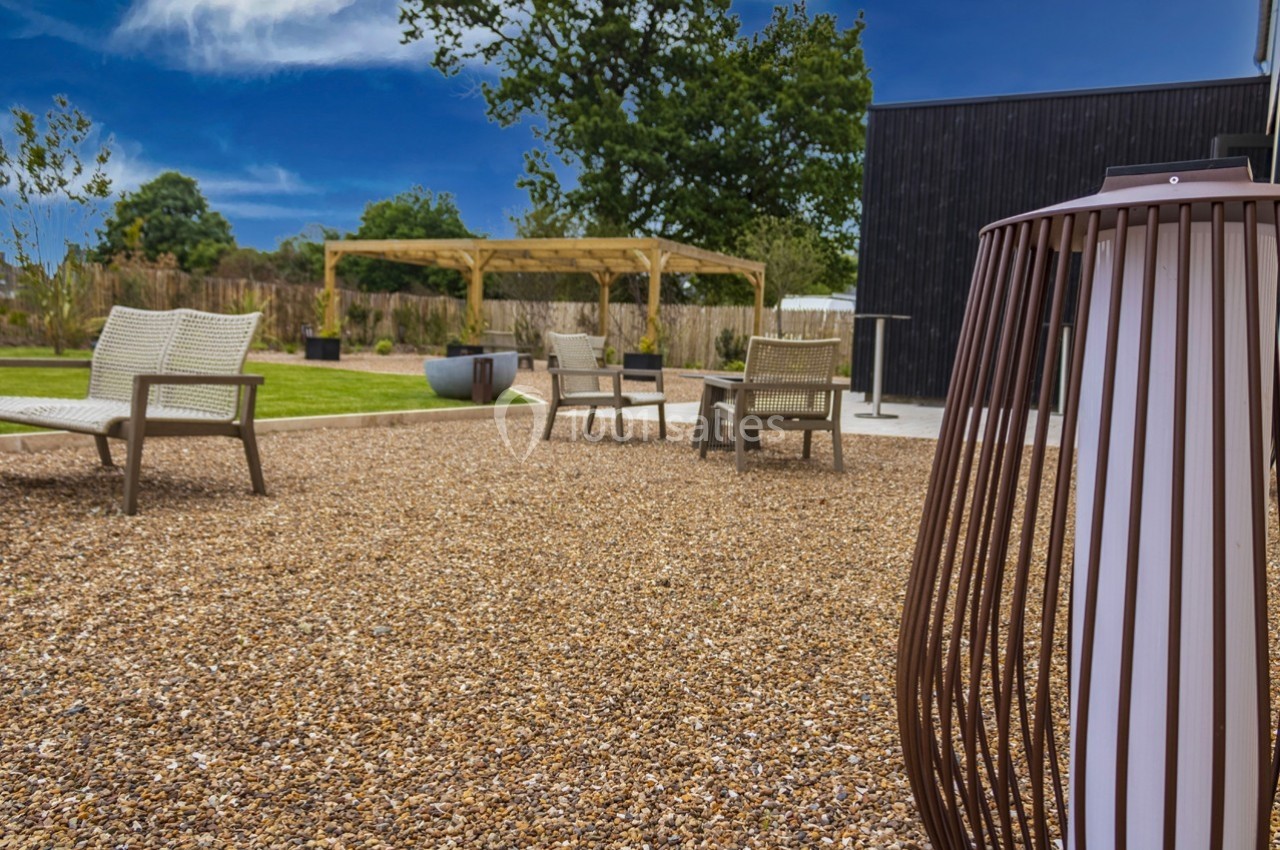 Espace extérieur avec gravier, fauteuils en osier, pergola en bois et végétation sous un ciel bleu.