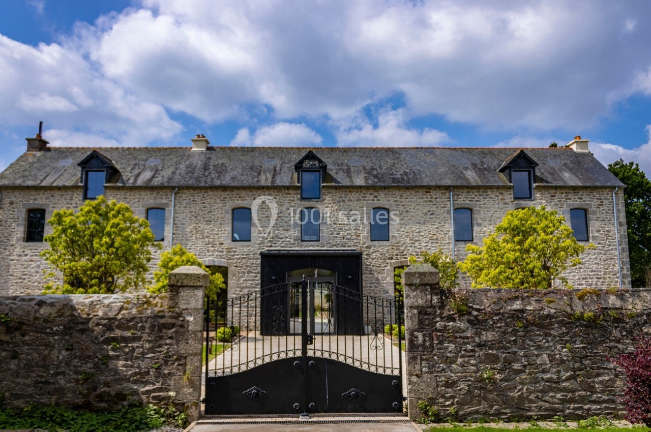 Façade d'une maison en pierre avec toit en ardoise, portail métallique noir et jardin arboré devant.