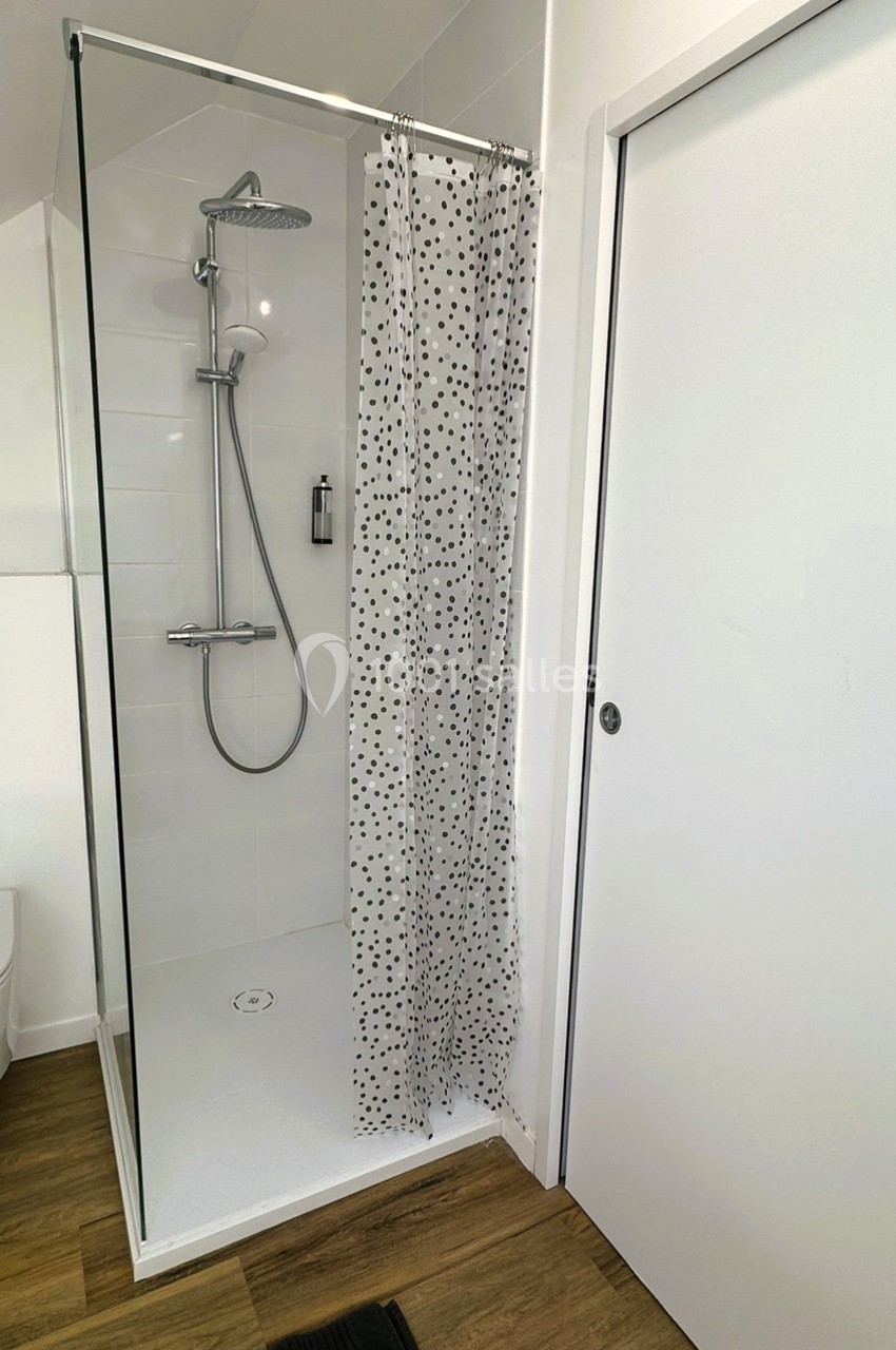 Douche moderne avec paroi vitrée, rideau à motifs noirs, sol en bois et porte blanche adjacente.