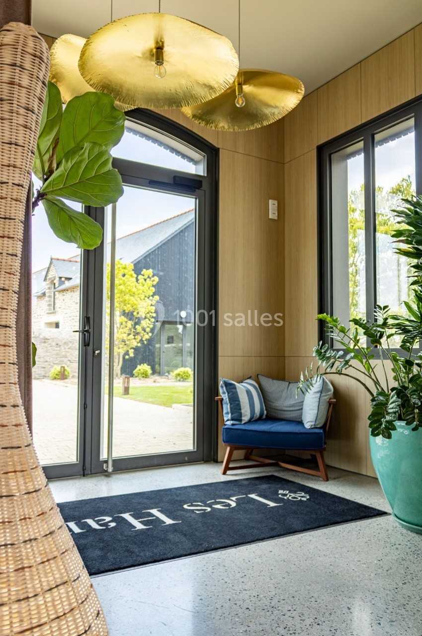 Entrée lumineuse avec fauteuil bleu, plantes en pot et tapis noir près d'une porte vitrée donnant sur l'extérieur.