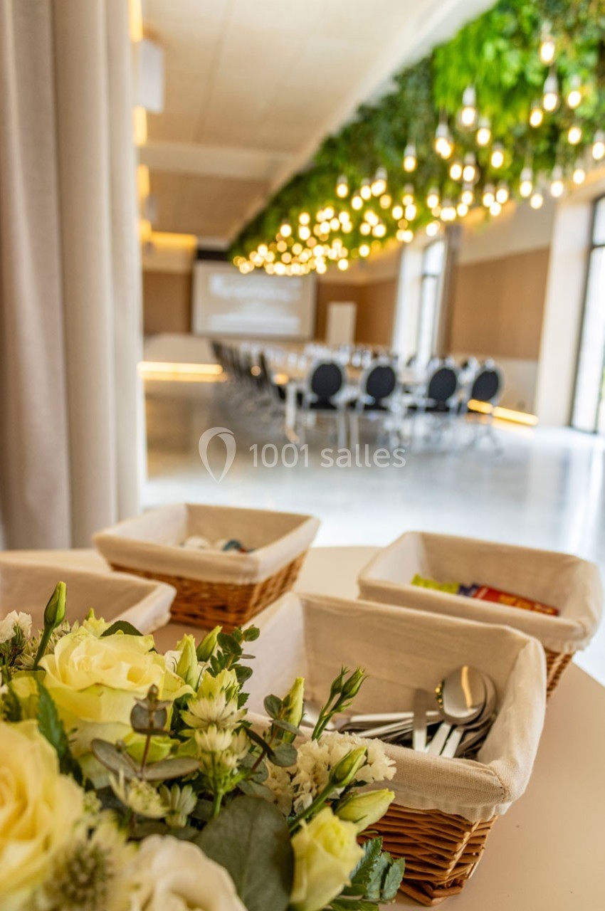 Salle de réunion lumineuse avec tables disposées en U, décorée de plantes suspendues et d'un bouquet au premier plan.