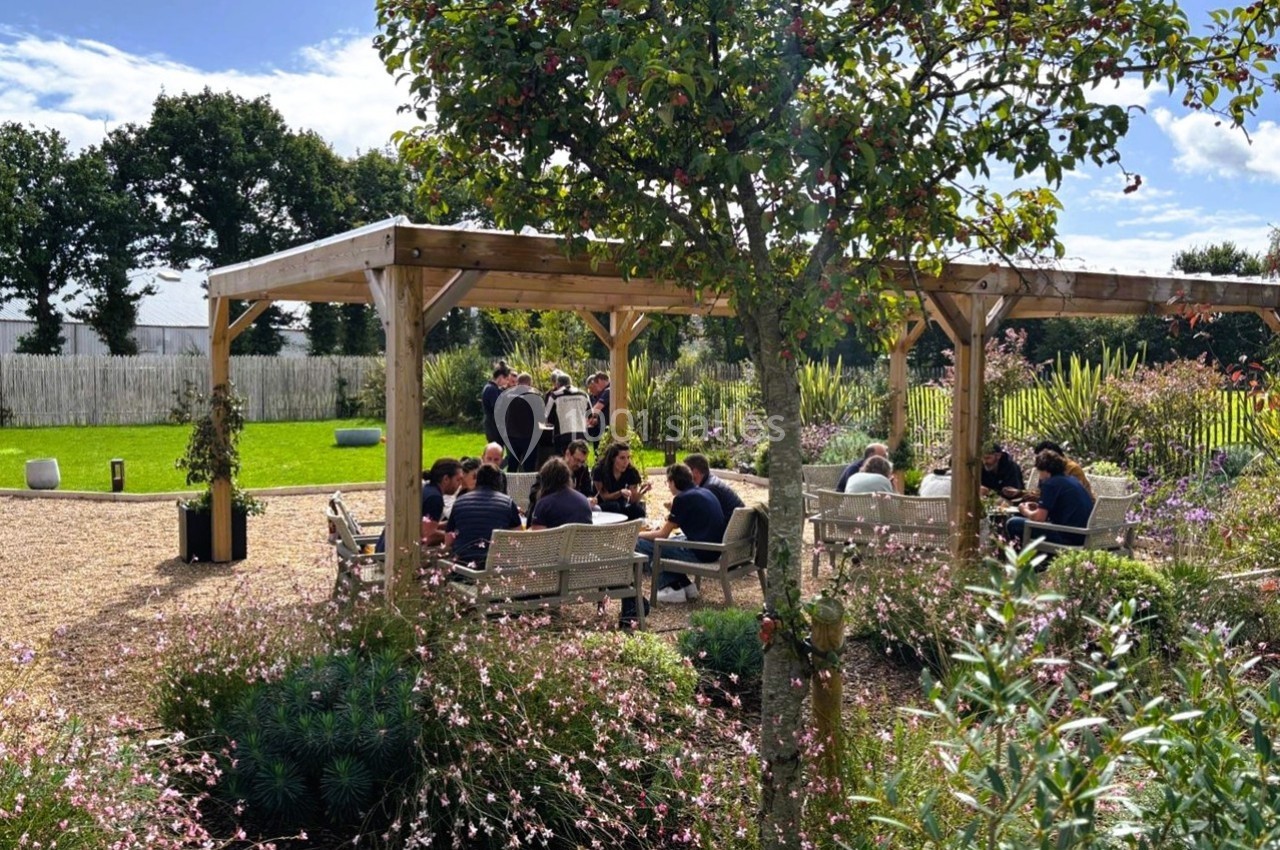 Des personnes assises sous une pergola en bois dans un jardin fleuri, entourées de verdure et de lumière naturelle.