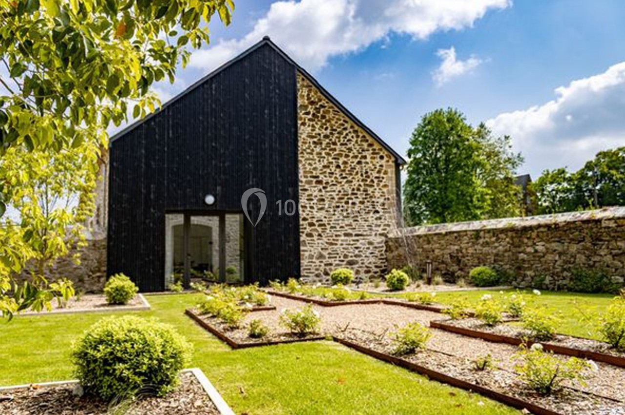 Façade d'une maison en pierre et bois noir, entourée d'un jardin paysager avec allées gravillonnées et arbustes.
