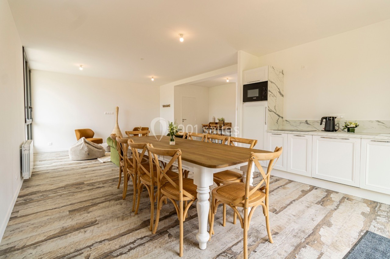 Salle à manger lumineuse avec une grande table en bois, des chaises en bois clair et une cuisine ouverte en arrière-plan.