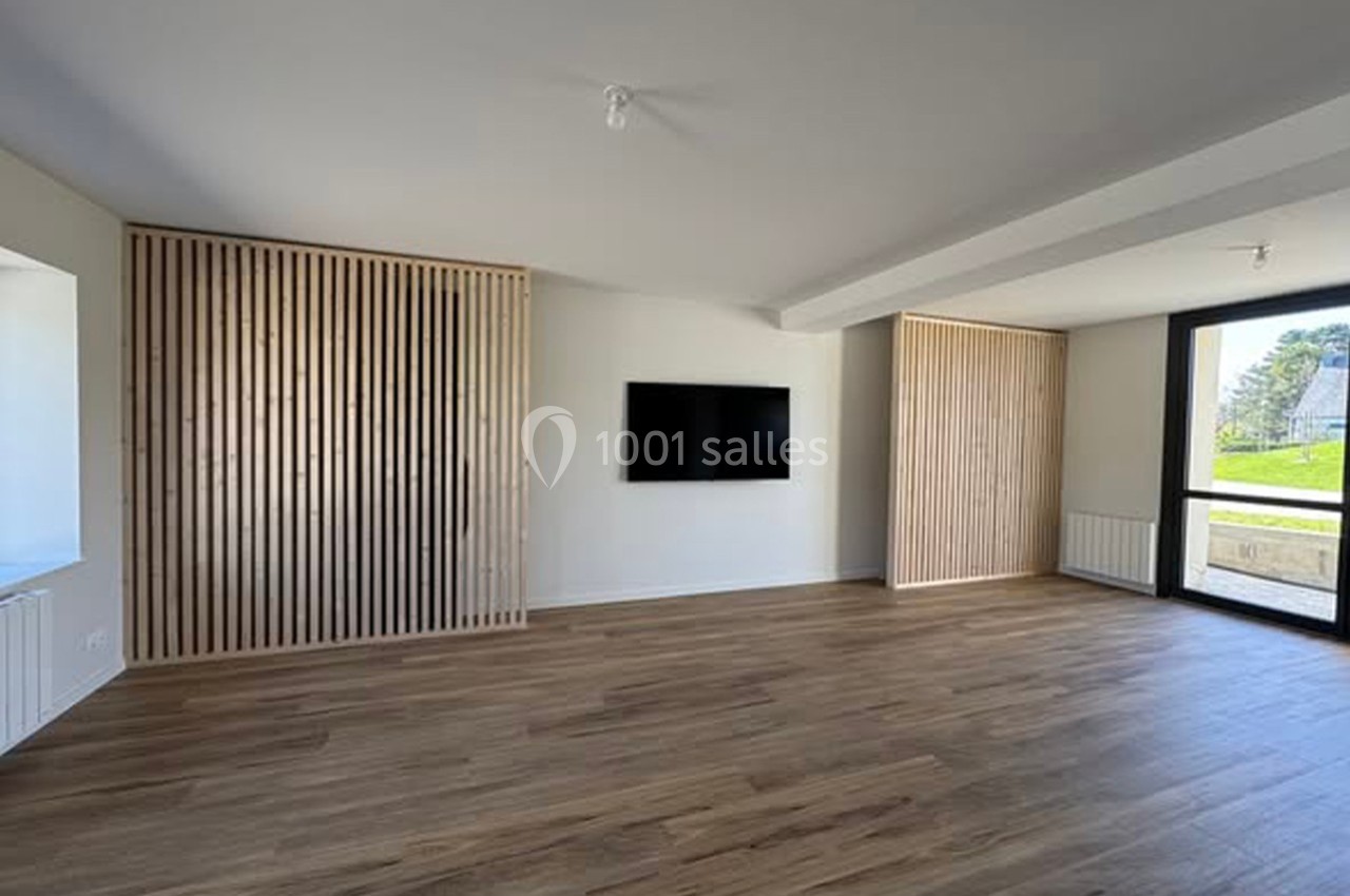 Pièce lumineuse avec parquet, murs blancs, panneaux en bois, télévision murale et grande fenêtre donnant sur l'extérieur.