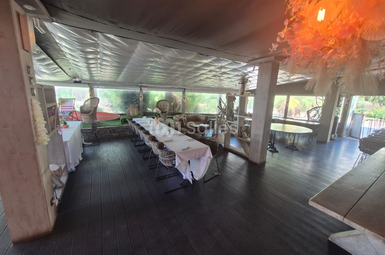 Salle de réception sous véranda avec table dressée, chaises en rotin et vue sur la végétation environnante.