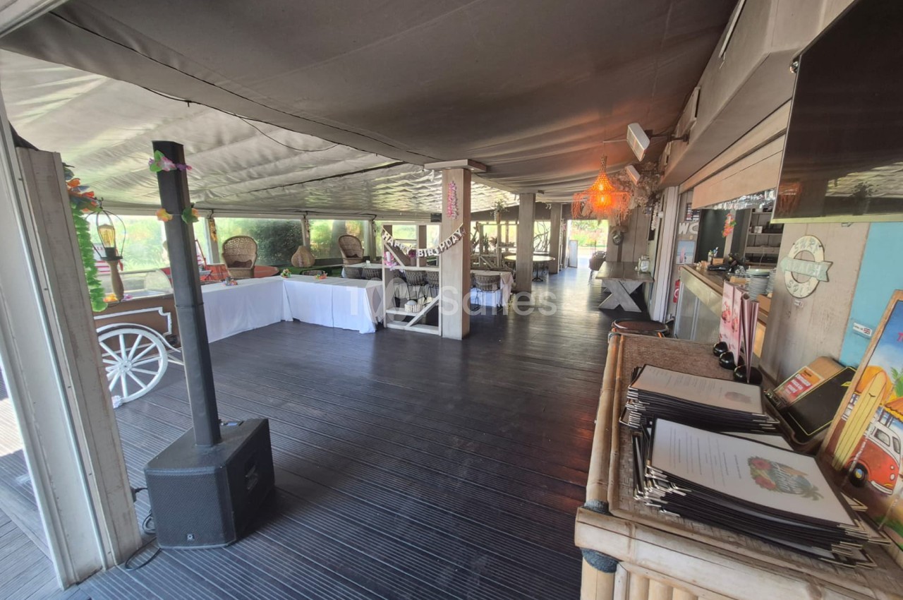 Salle couverte avec sol en bois, comptoir blanc, chaises en rotin et baies vitrées donnant sur jardin.