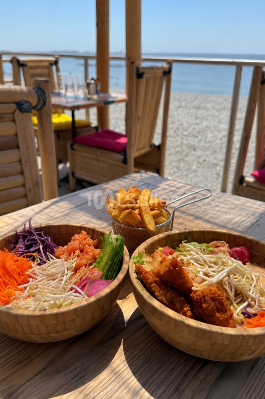 Terrasse bord de mer avec deux bols de nouilles, fried chicken et corbeille de frites, vue sur plage et mer.