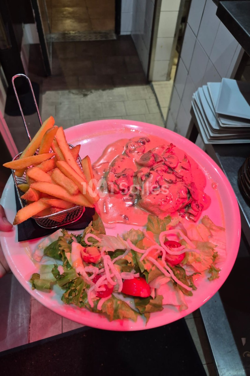 Assiette rose contenant des frites, de la viande rouge hachée et une salade verte, en cuisine professionnelle.