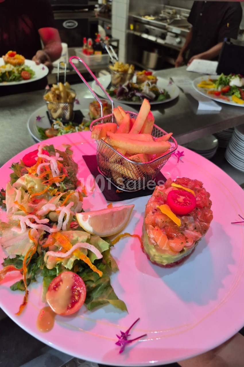 Assiette rose avec tartare de poisson, frites en panier métallique rose et légumes colorés en arrière-plan en cuisine.