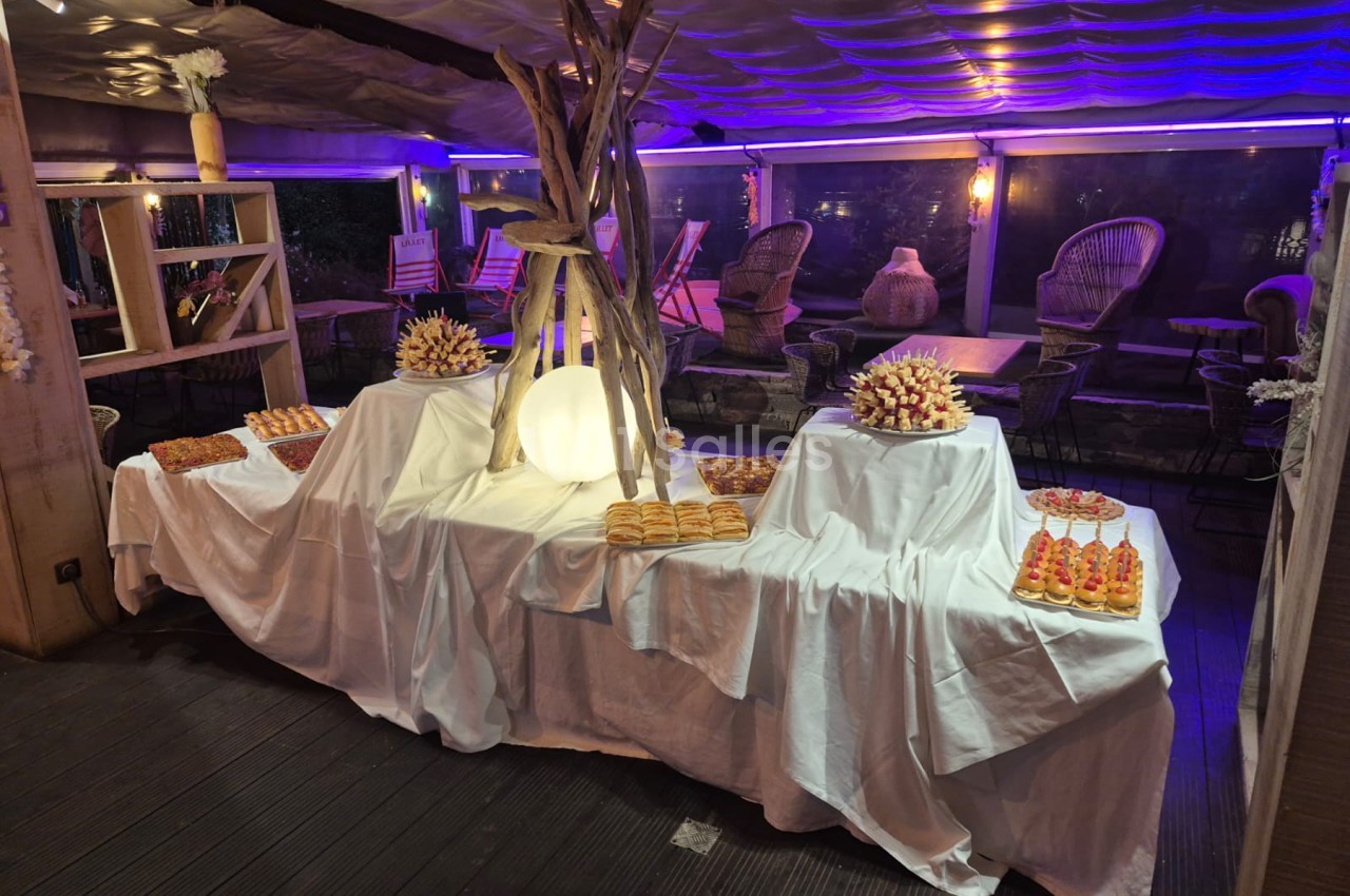 Salle intérieure aux éclairages bleus et violets, tables dressées avec nappes blanches présentant des pâtisseries et compositions florales.