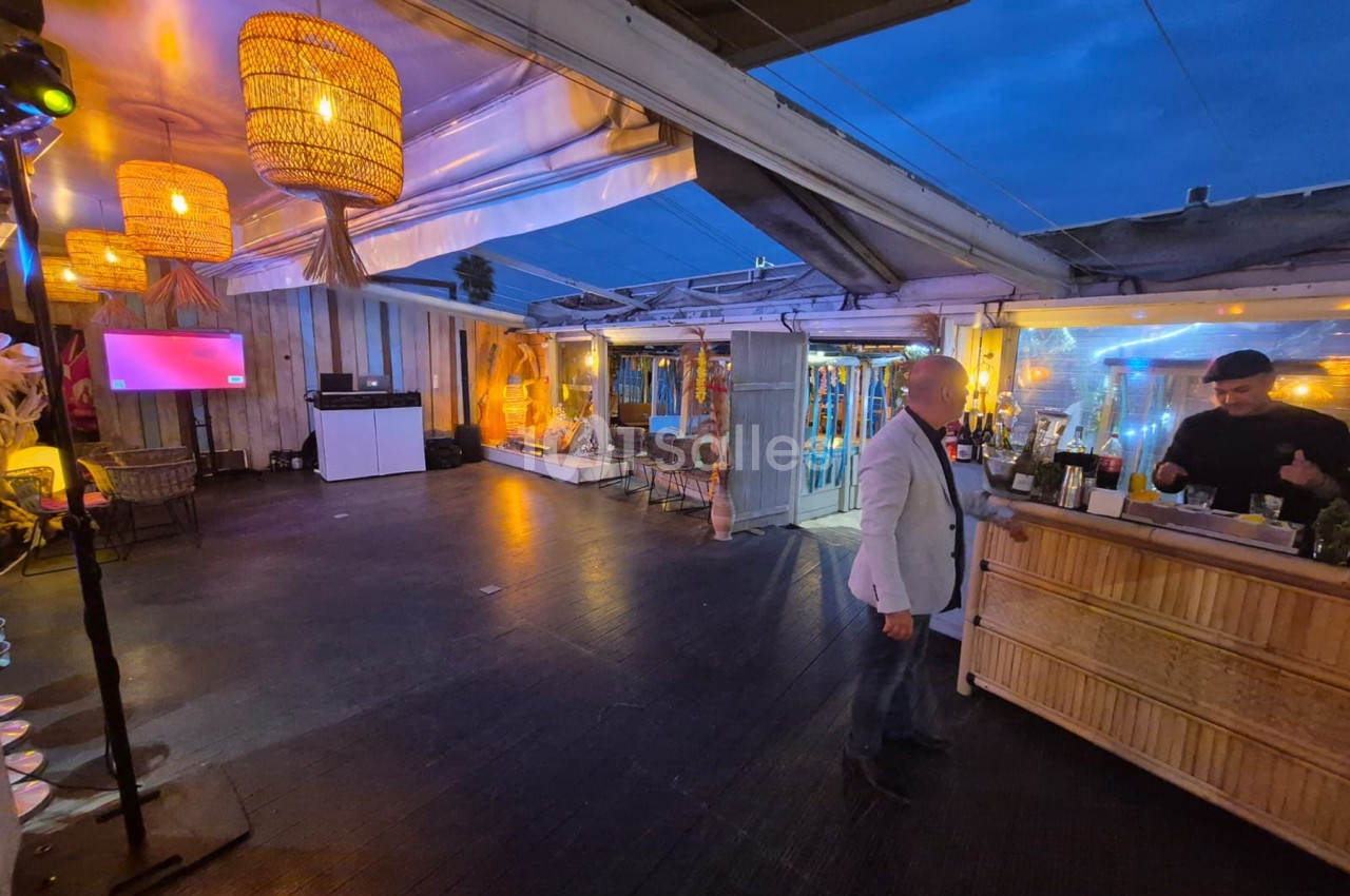 Salle de réception avec bar à droite, piste de danse centrale, luminaires en rotin suspendus, éclairage jaune et rose, plafond vitré dégageant vue nocturne.