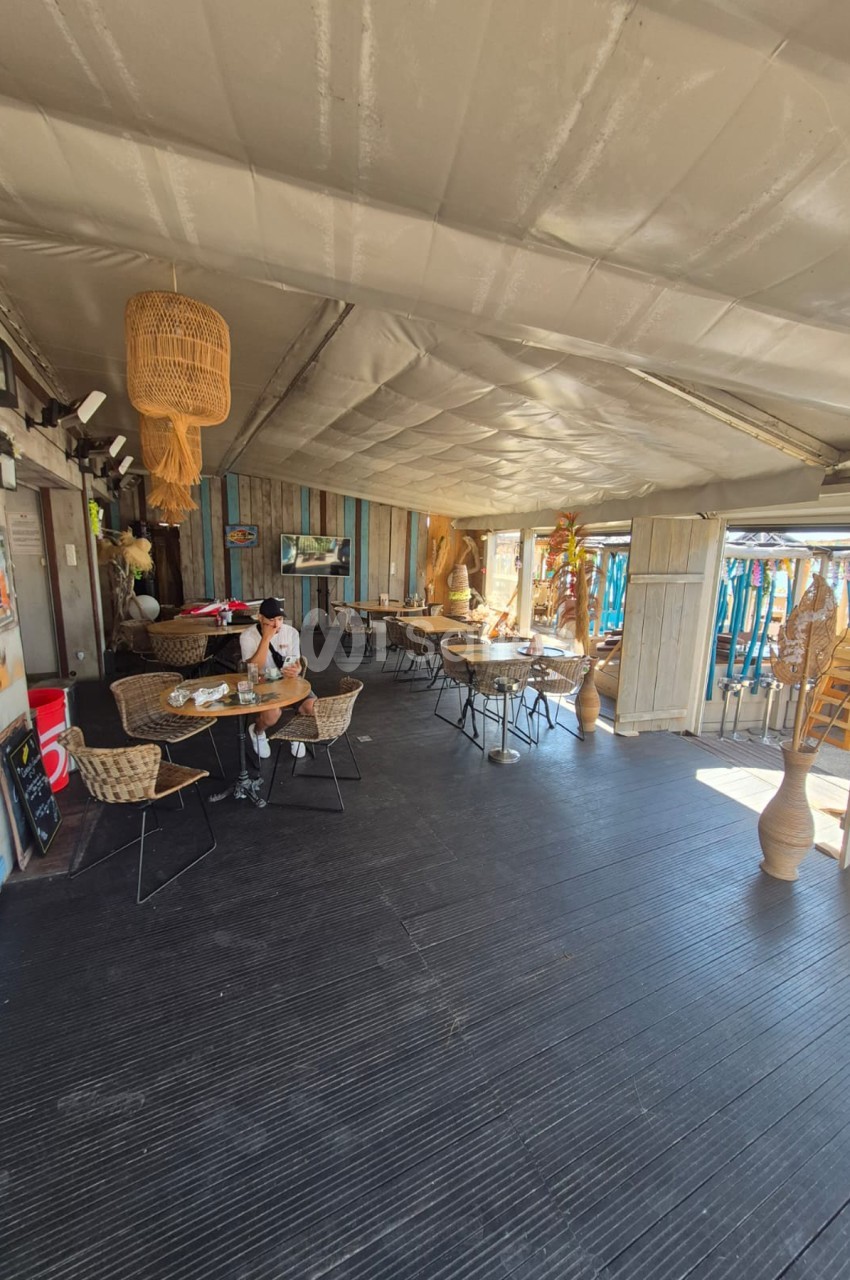 Intérieur spacieux avec plafond blanc tendu, sol gris foncé, tables basses en rotin, chaises noires, suspensions en osier et décoration bohème - une personne attablée à gauche.
