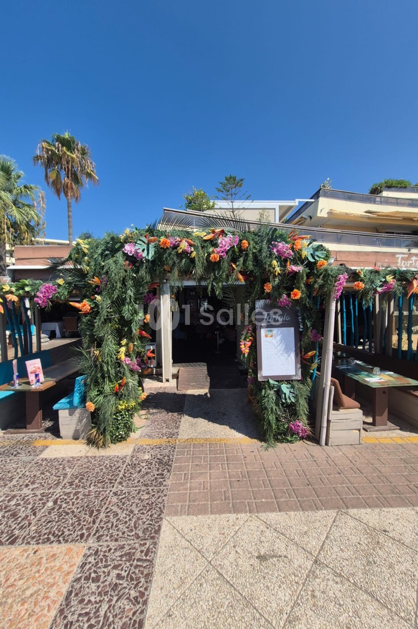 Entrée extérieure d'un restaurant décorée d'une arche florale rose et orange sous un ciel bleu dégagé.
