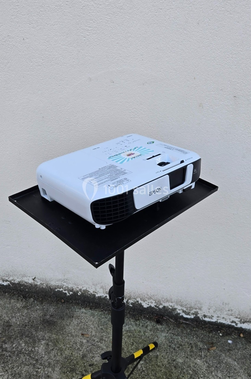 Projecteur blanc posé sur un support noir, devant un mur clair et un sol en béton.