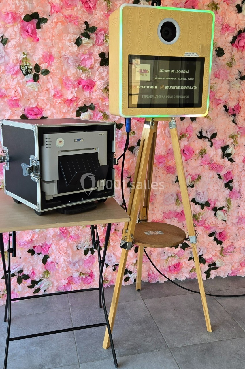Borne photo en bois sur trépied avec imprimante à côté, devant un mur décoré de fleurs roses.