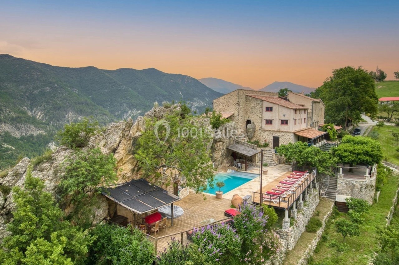 Vue aérienne d'une maison en pierre avec piscine, terrasse et transats, entourée de montagnes et de végétation.