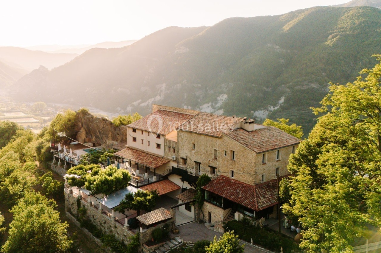 Vue aérienne d'un bâtiment en pierre entouré de verdure, situé dans une vallée montagneuse au coucher du soleil.