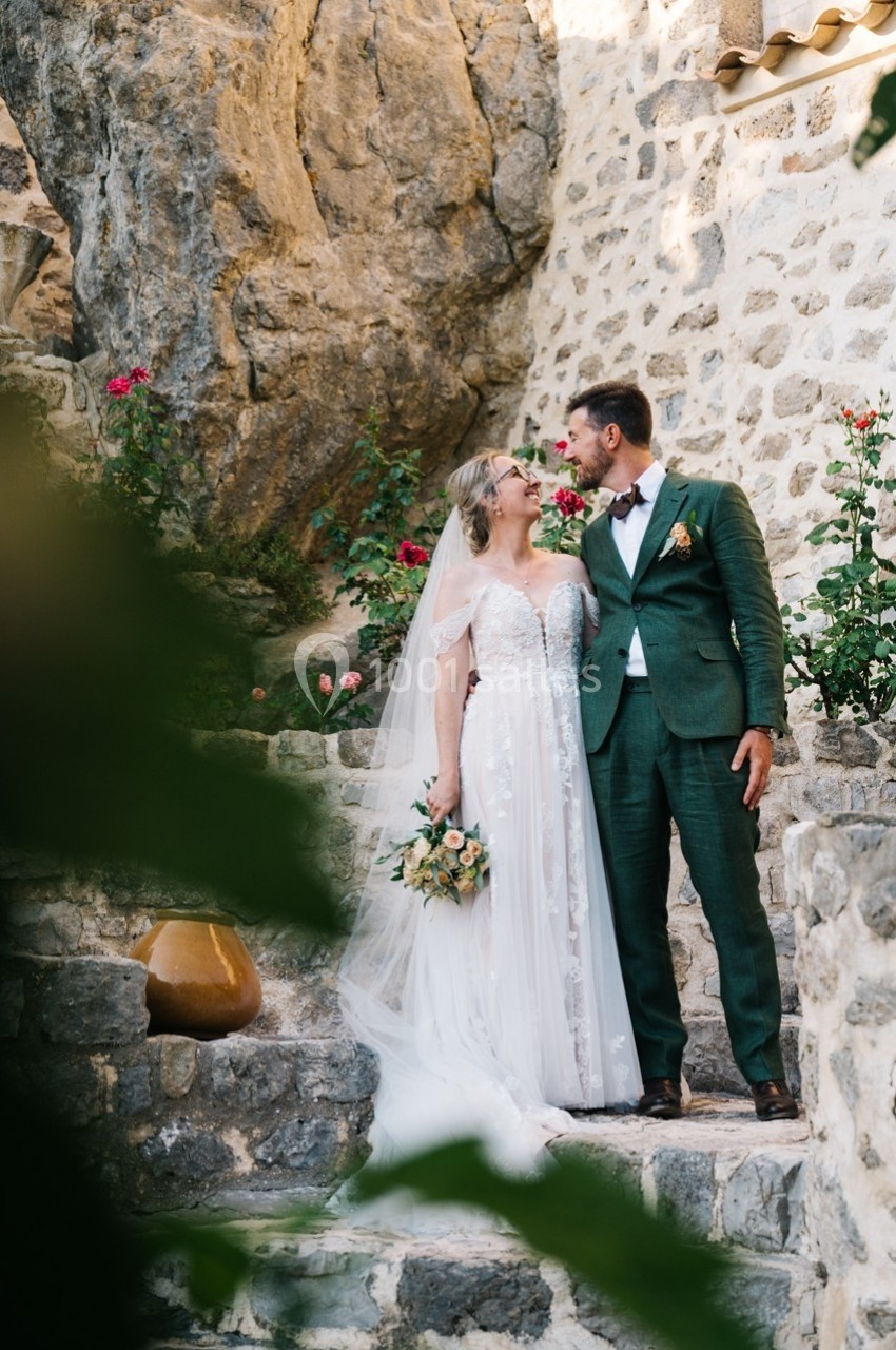 Un couple de mariés souriant se tient sur des marches en pierre, entouré de fleurs et de murs en pierre.