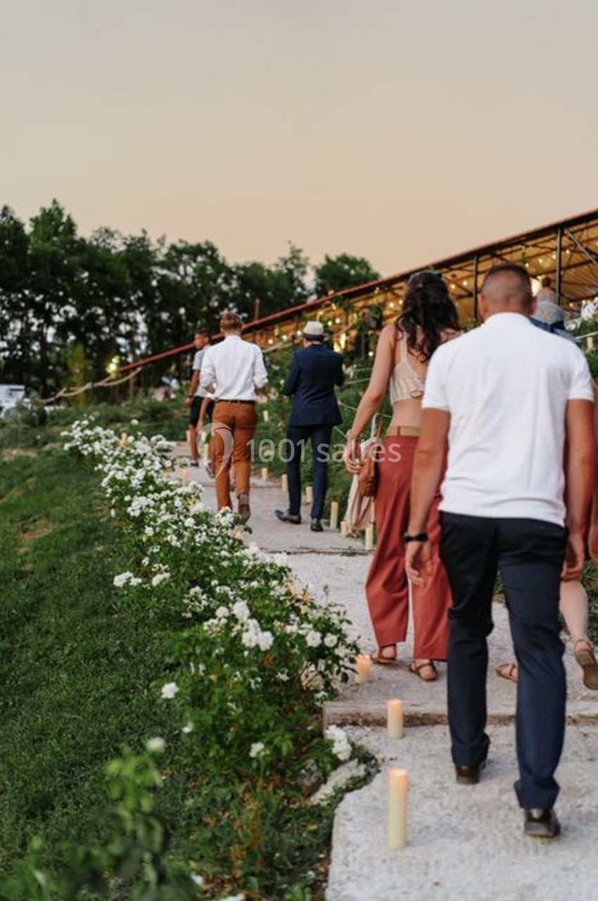 Des personnes marchent sur un chemin bordé de fleurs blanches et de bougies, en extérieur au crépuscule.