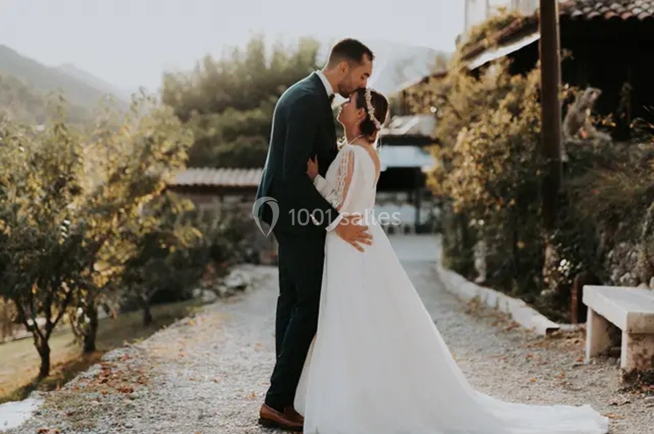 Un couple en tenue de mariage s'embrasse tendrement dans une allée bordée de verdure, sous une lumière douce.