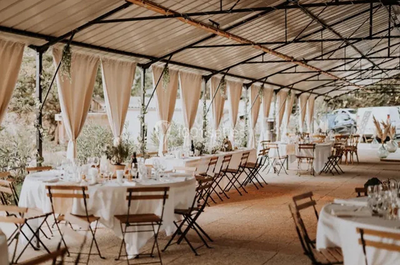 Salle extérieure couverte avec tables et chaises disposées pour un événement, entourée de rideaux et de verdure.