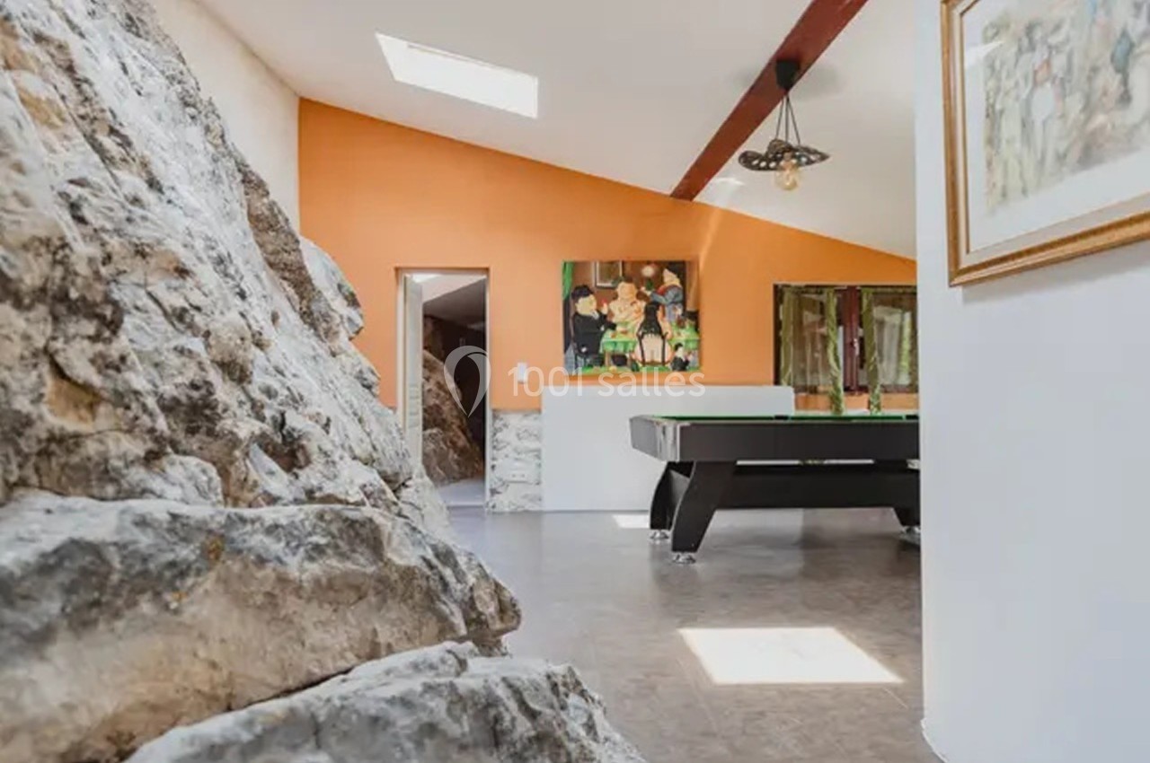 Salle avec un mur en pierre naturelle, un billard noir, un mur orange et une lumière naturelle provenant d'une lucarne.