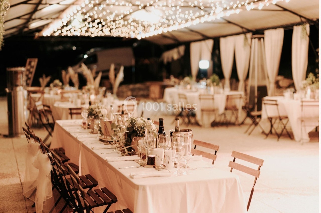Salle de réception en extérieur avec tables décorées, chaises en bois et guirlandes lumineuses suspendues.