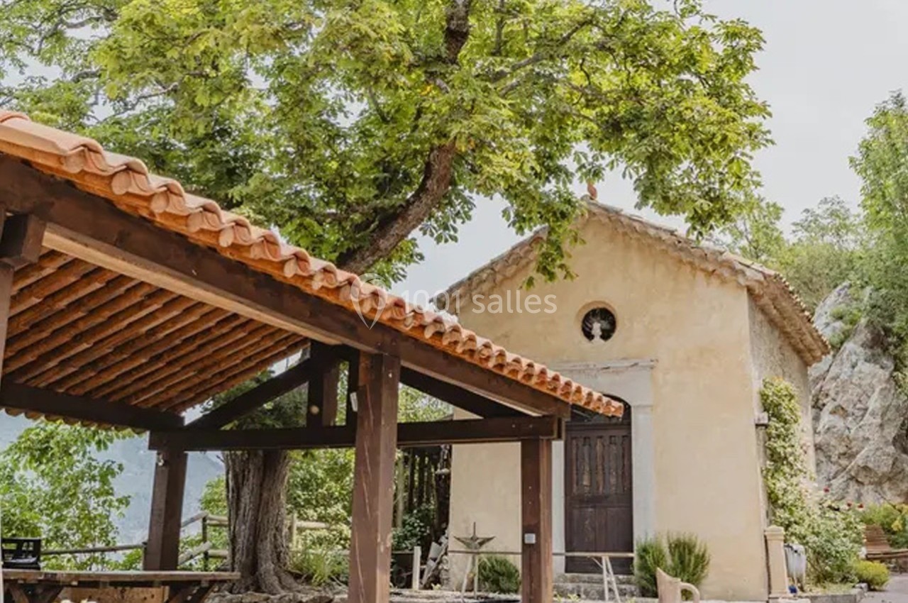 Petite chapelle en pierre avec toit en tuiles, entourée de verdure et d'une pergola en bois au premier plan.