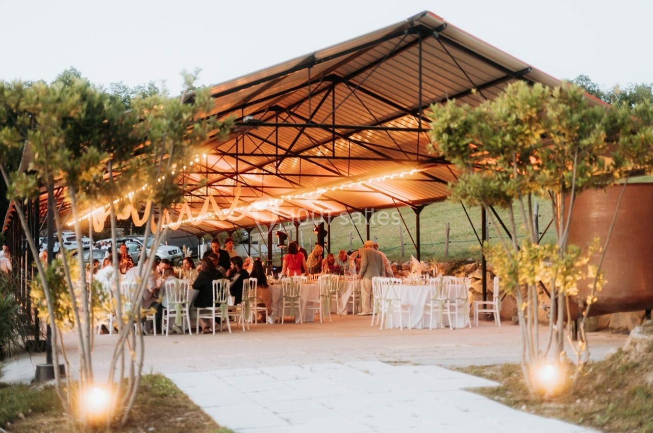 Salle de réception en plein air sous une structure métallique, décorée de guirlandes lumineuses, avec des invités assis.