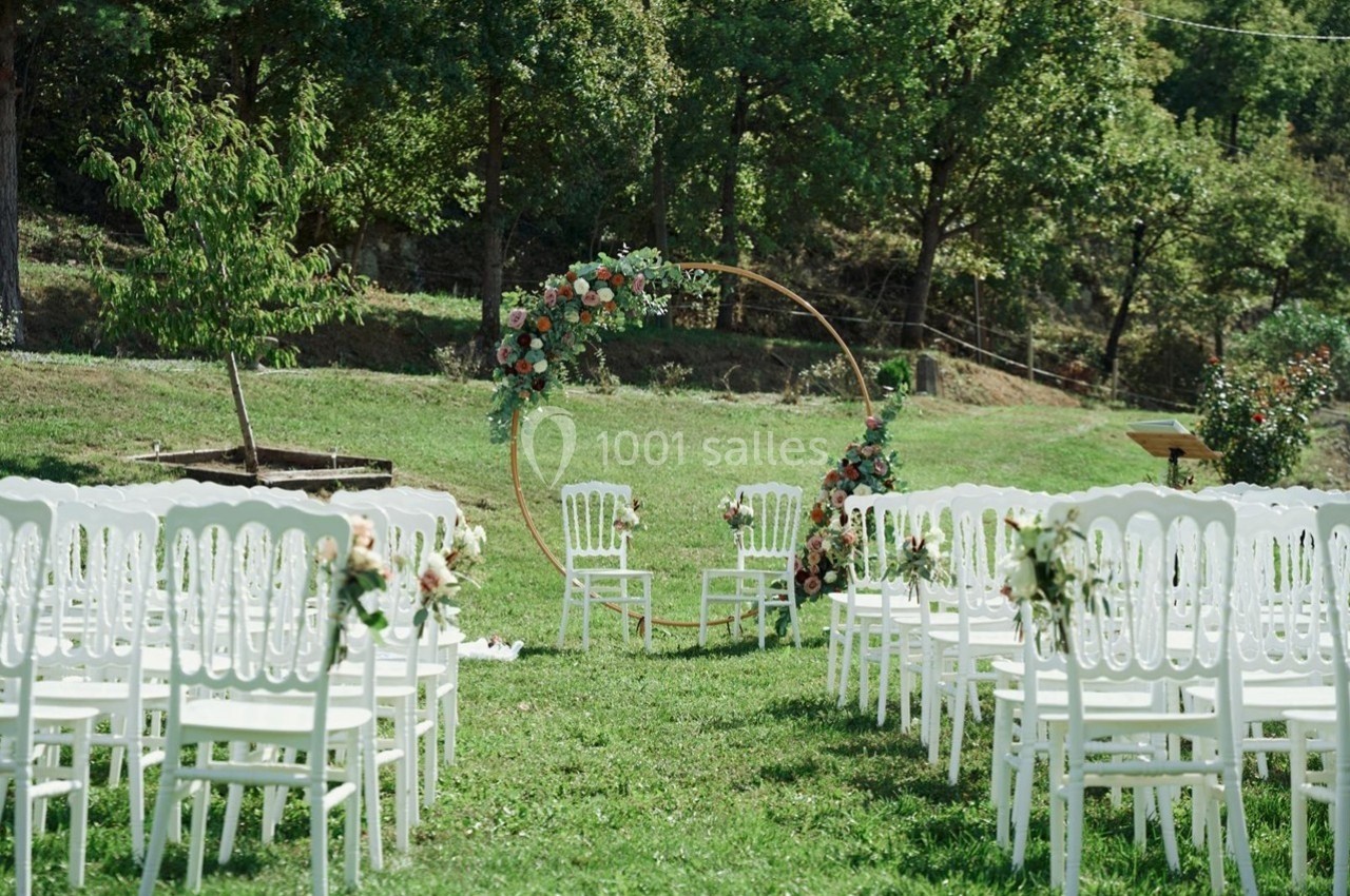 Chaises blanches disposées en allée face à une arche florale circulaire dans un jardin verdoyant.