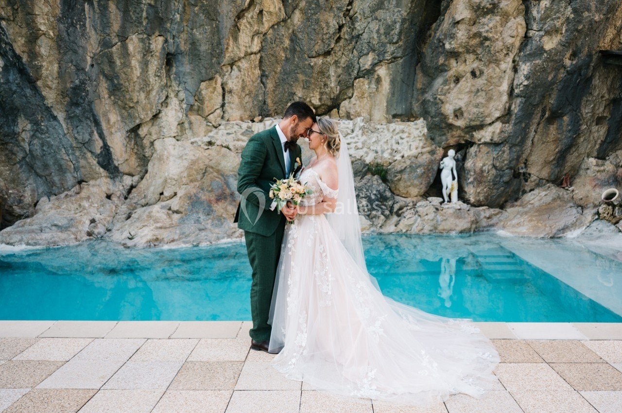 Un couple de mariés échange un regard tendre devant une piscine et une paroi rocheuse.
