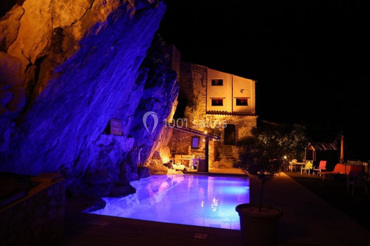 Piscine éclairée en bleu la nuit, entourée de rochers et d'une maison en pierre avec des lumières chaleureuses.
