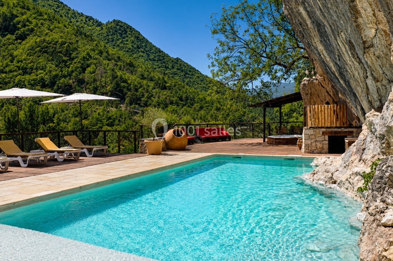 Piscine extérieure entourée de montagnes verdoyantes, avec chaises longues, parasols et terrasse en pierre.