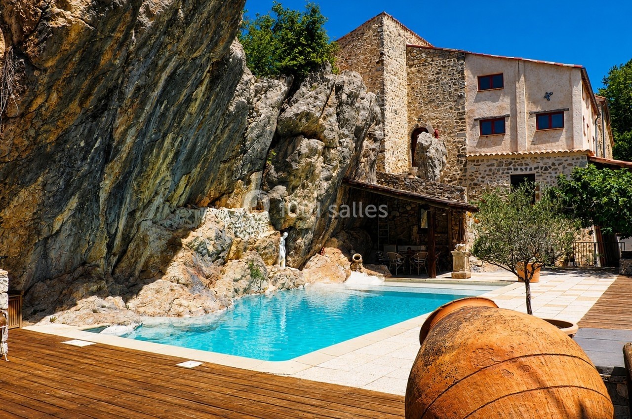 Piscine extérieure entourée de rochers, terrasse en bois et maison en pierre sous un ciel bleu.