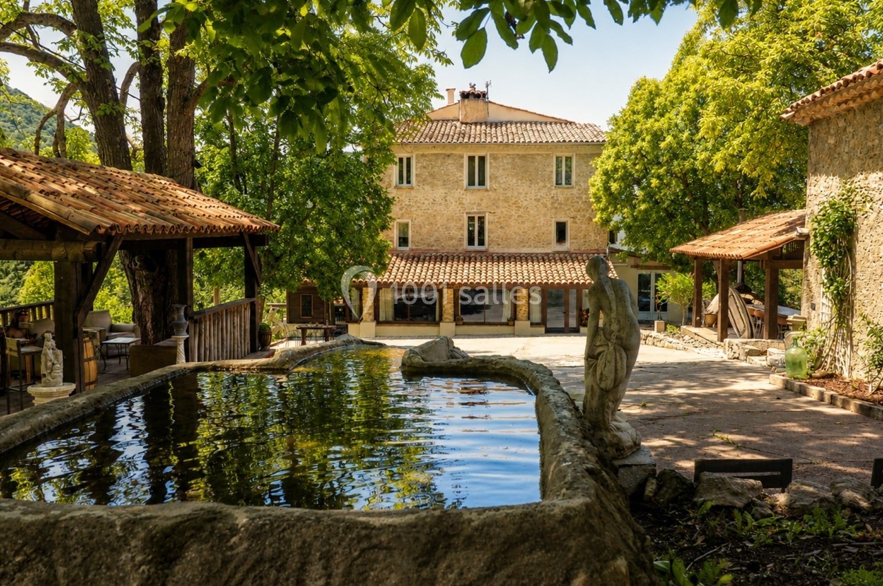 Cour d'une maison en pierre entourée d'arbres, avec un bassin en pierre et une statue au premier plan.