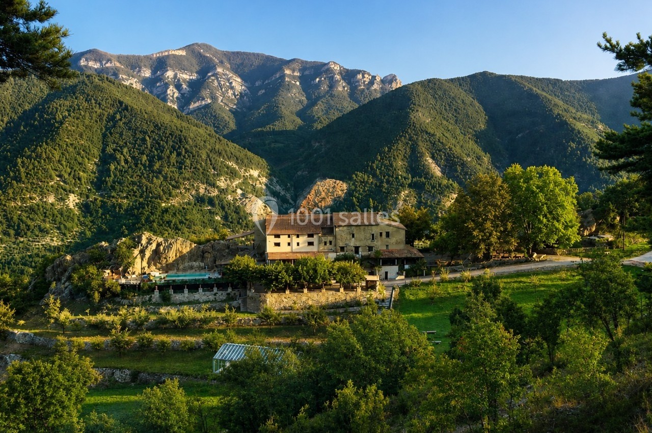 Village rural entouré de montagnes verdoyantes sous un ciel bleu clair, avec des arbres et des champs au premier plan.