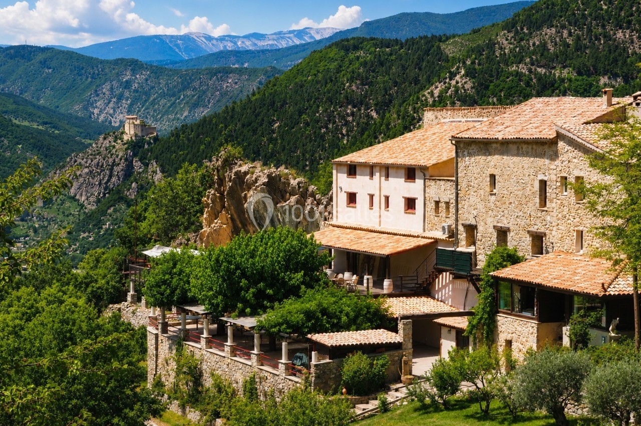 Vue d'un village en pierre entouré de verdure et de montagnes sous un ciel bleu.