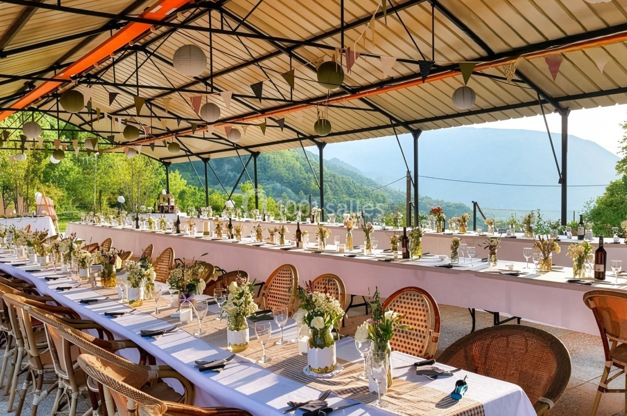 Tables décorées pour un repas en plein air sous un auvent, avec vue sur des collines verdoyantes en arrière-plan.