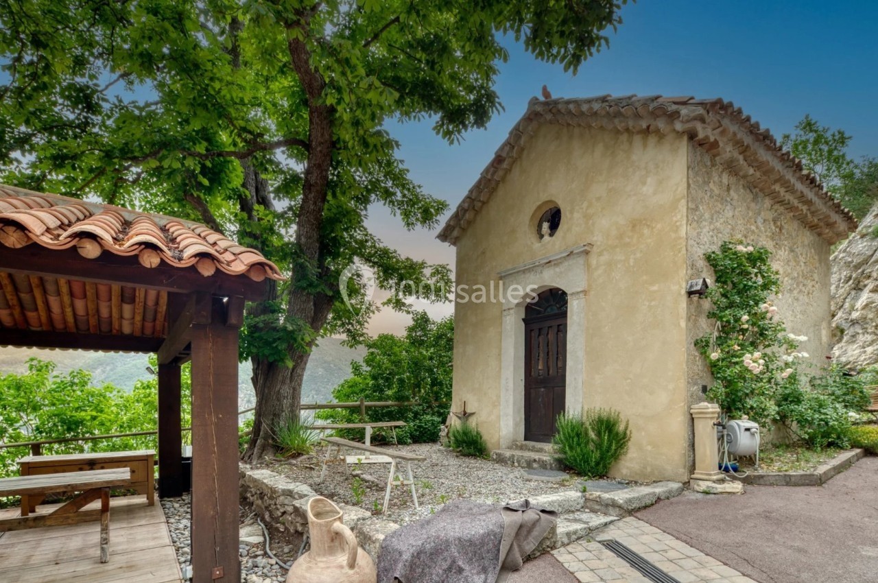 Petite chapelle en pierre entourée de végétation, avec un arbre et une terrasse couverte en bois à proximité.