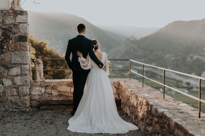 Miniature Location salle Entrevaux (Alpes-de-Haute-Provence) - Domaine de Glandève #14 Un couple en tenue de mariage regarde un paysage montagneux au coucher du soleil depuis une terrasse en pierre.