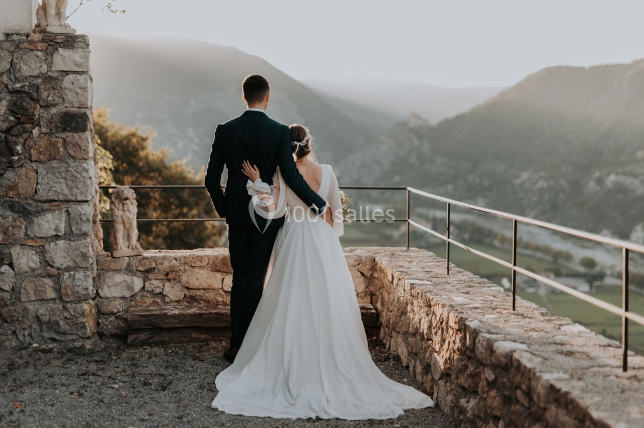 Un couple en tenue de mariage regarde un paysage montagneux au coucher du soleil depuis une terrasse en pierre.