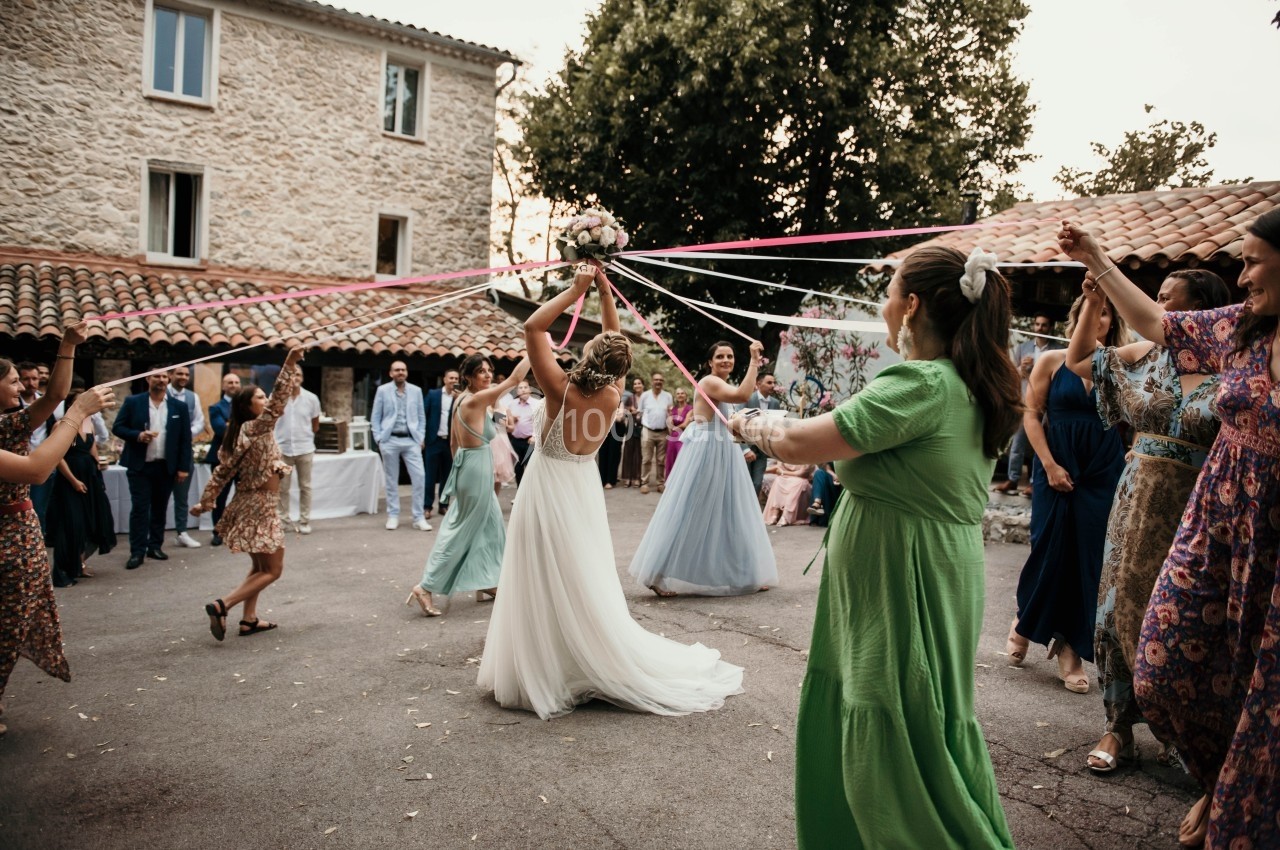 Des invités dansent autour d'une mariée tenant un bouquet, avec des rubans colorés, dans une cour extérieure.