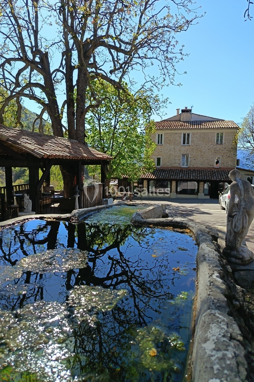 Bassin en pierre reflétant des arbres, avec une statue et une maison en pierre en arrière-plan sous un ciel clair.