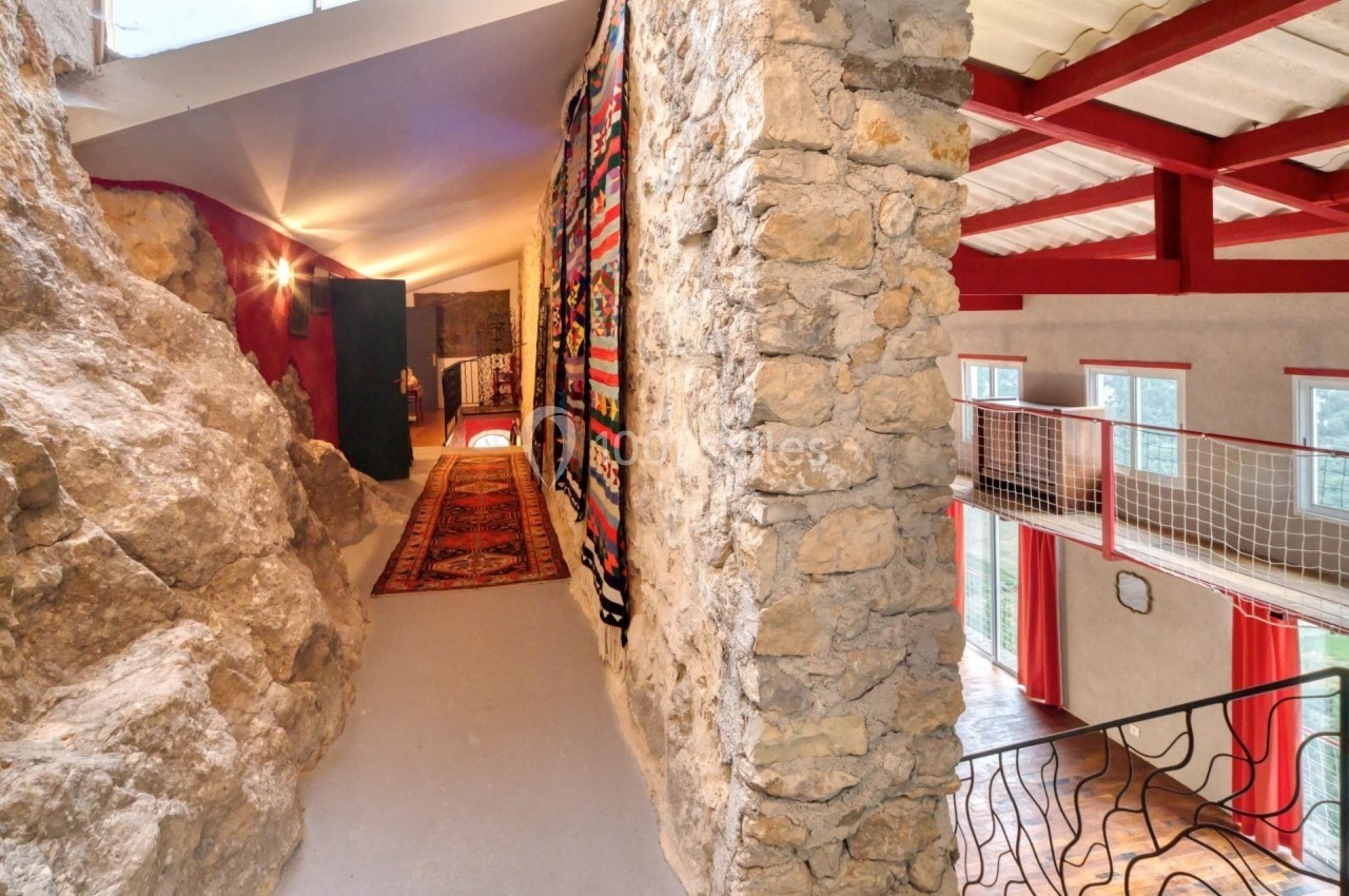 Couloir intérieur avec murs en pierre, tapis coloré au sol et vue sur une mezzanine lumineuse avec poutres rouges.