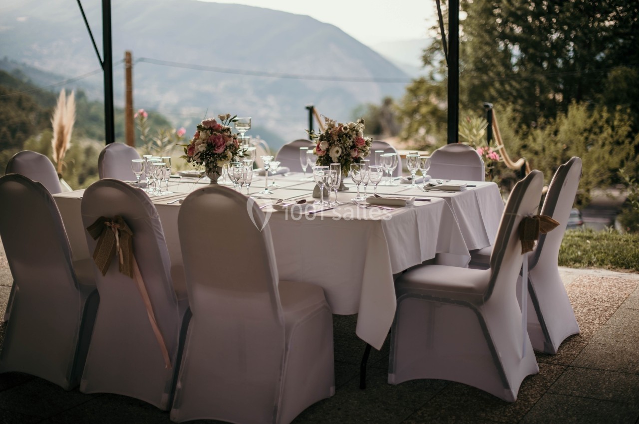Table dressée en extérieur avec nappes blanches, chaises couvertes et bouquets de fleurs, vue sur des collines en arrière…